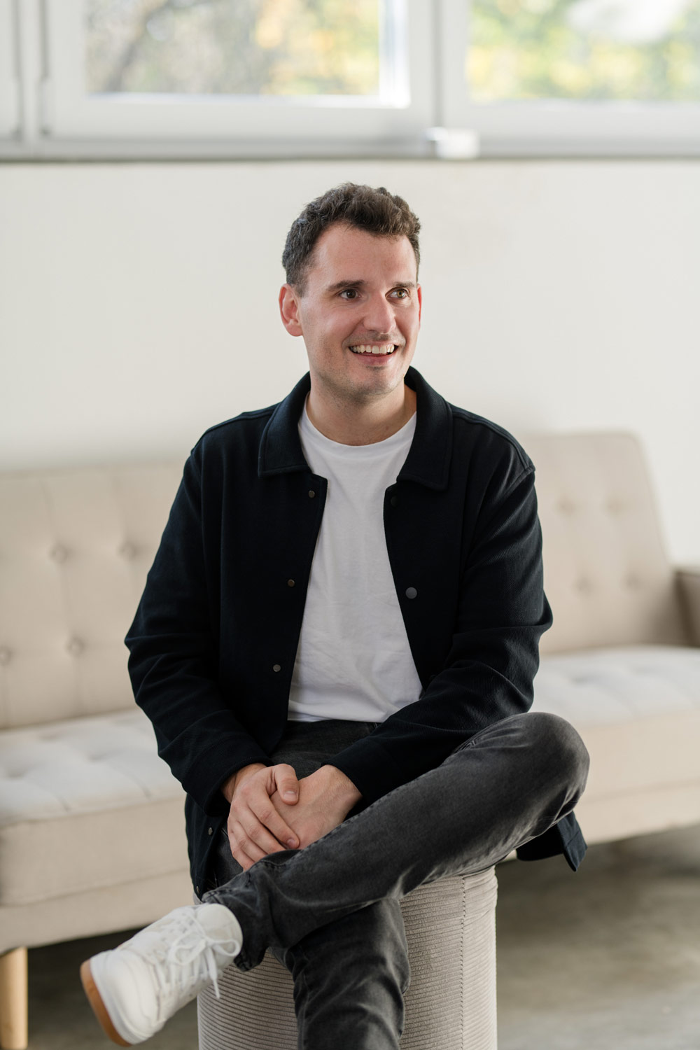 Jan-Christopher Gent sitzend in schwarzer Jacke und weißem T-Shirt, lächelnd